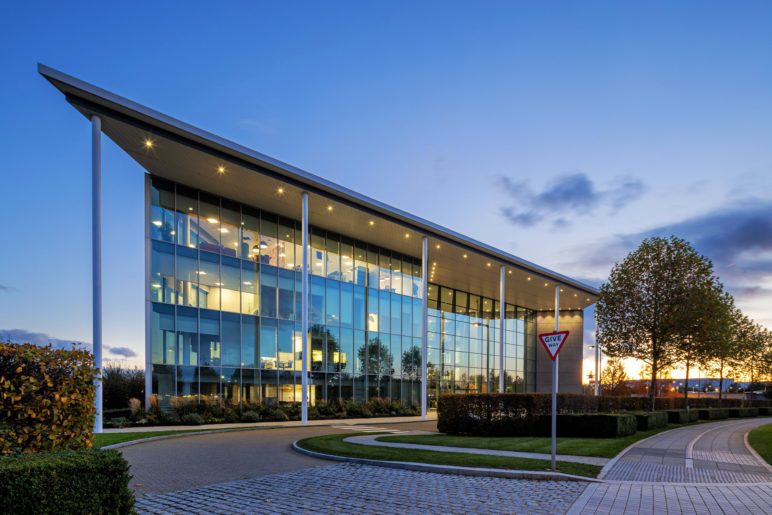 A modern glass office building at sunset features a curved driveway and landscaped surroundings