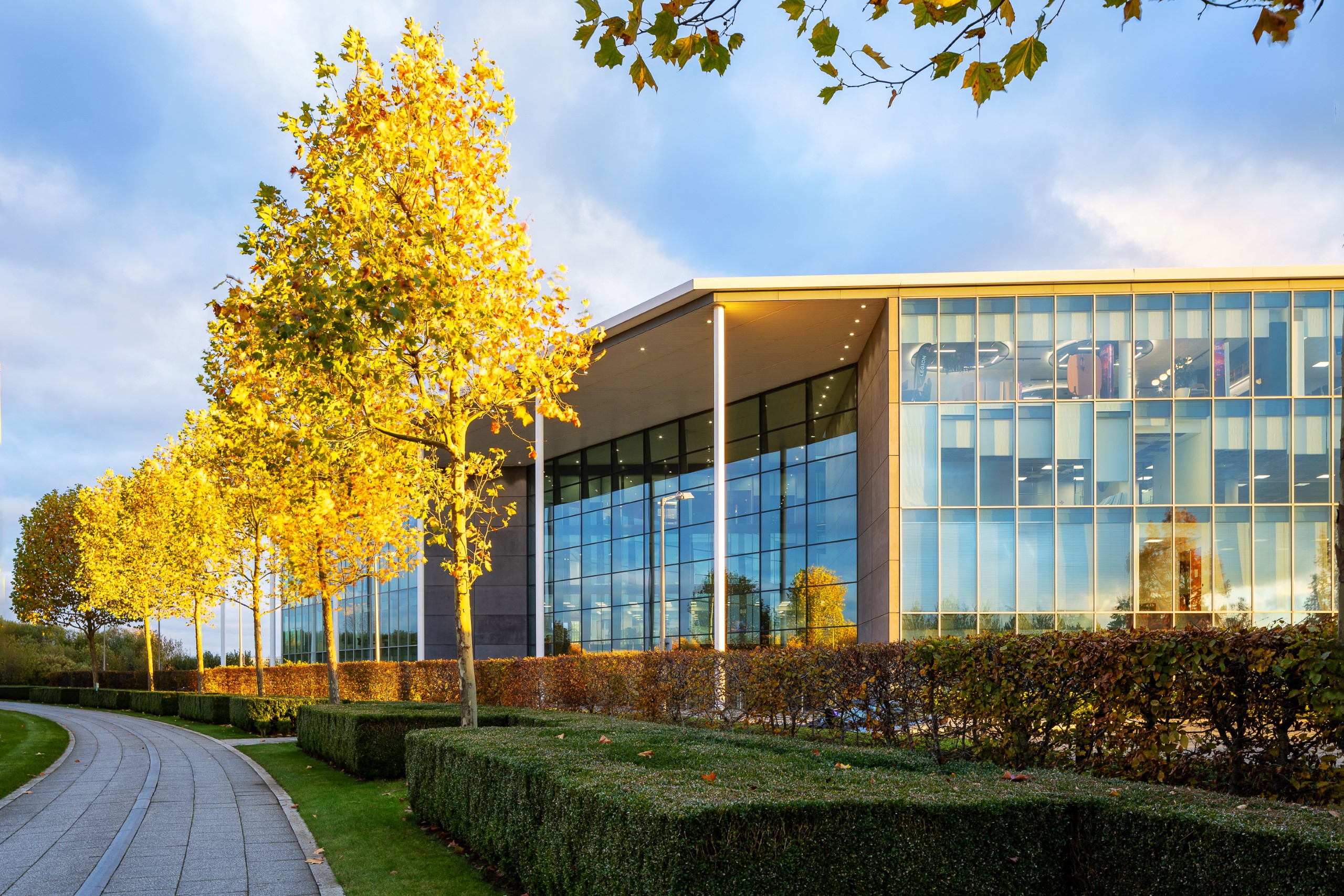 A modern office building with trees and hedges lining a curved walkway under a partly cloudy sky