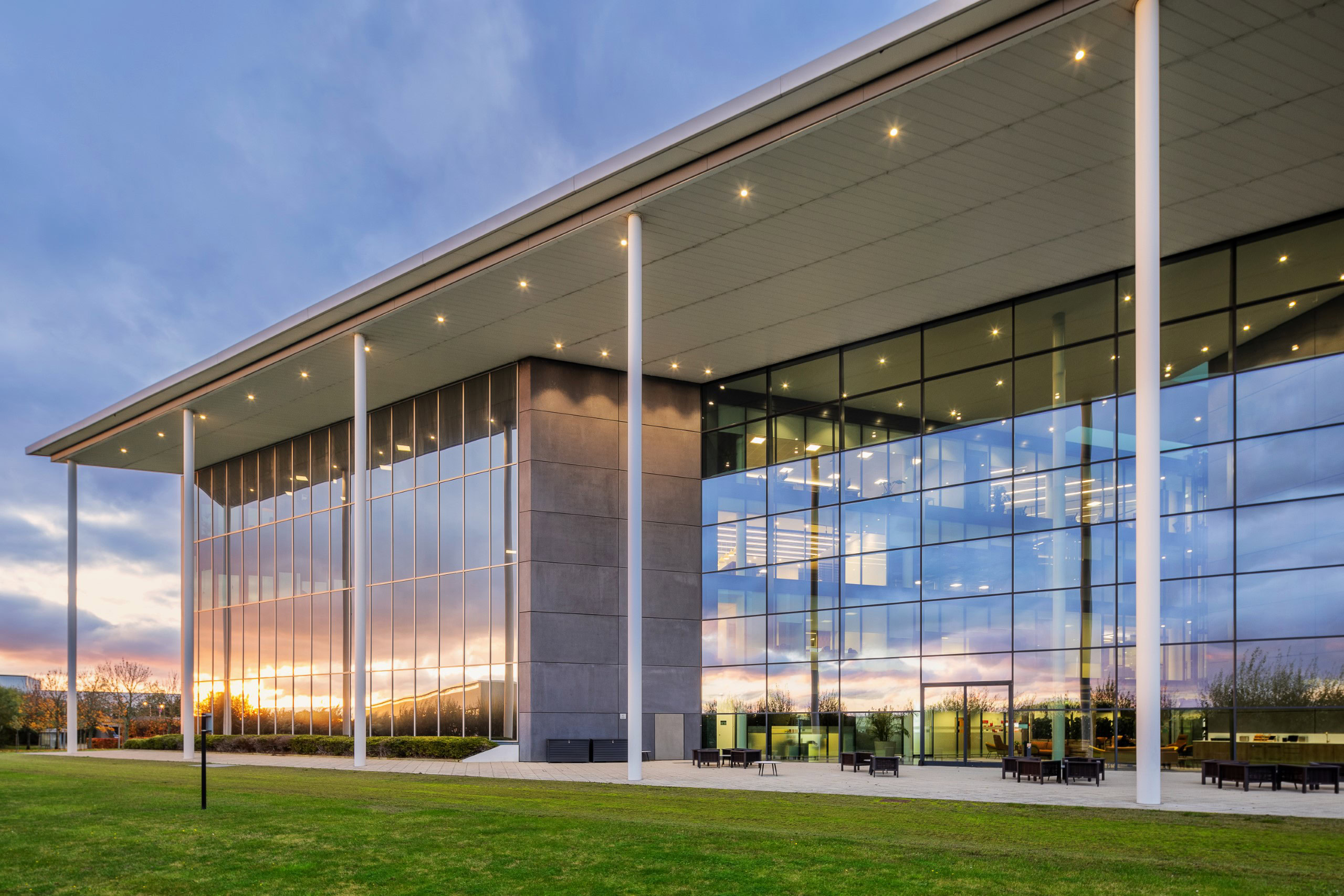 A modern glass & steel office building with a large overhanging roof reflects a colourful sunset sky