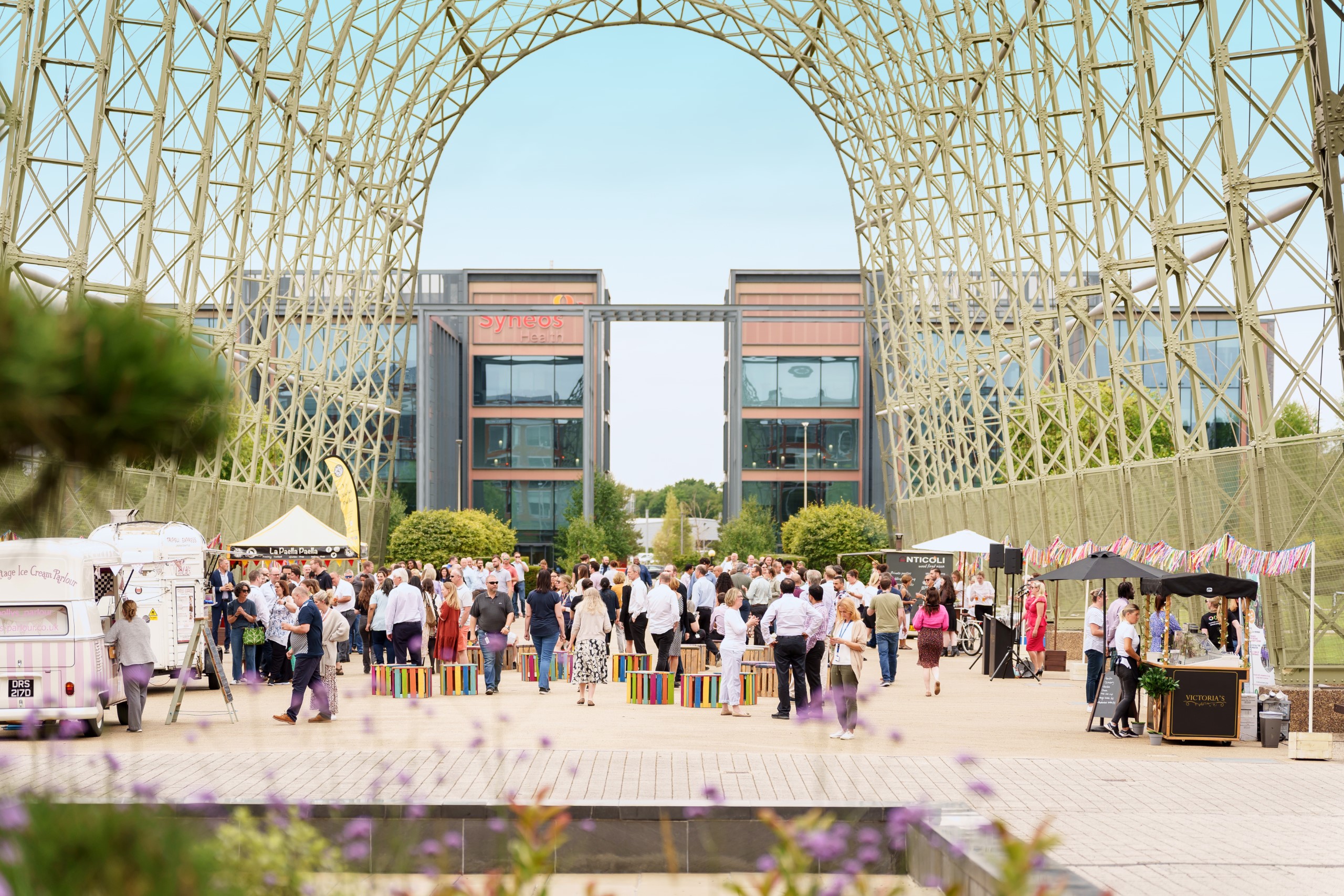 A large group of people gathers under a spacious metal canopy for an event. Food trucks and colorful decorations add vibrancy, set against the backdrop of an office building and lush greenery, making it a lively scene in the heart of a bustling business park.