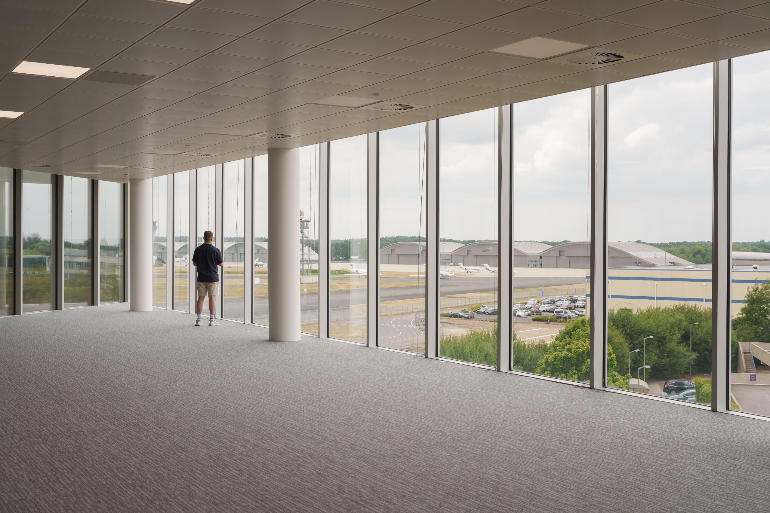A person stands in an office space with windows, overlooking an airport runway and hangars