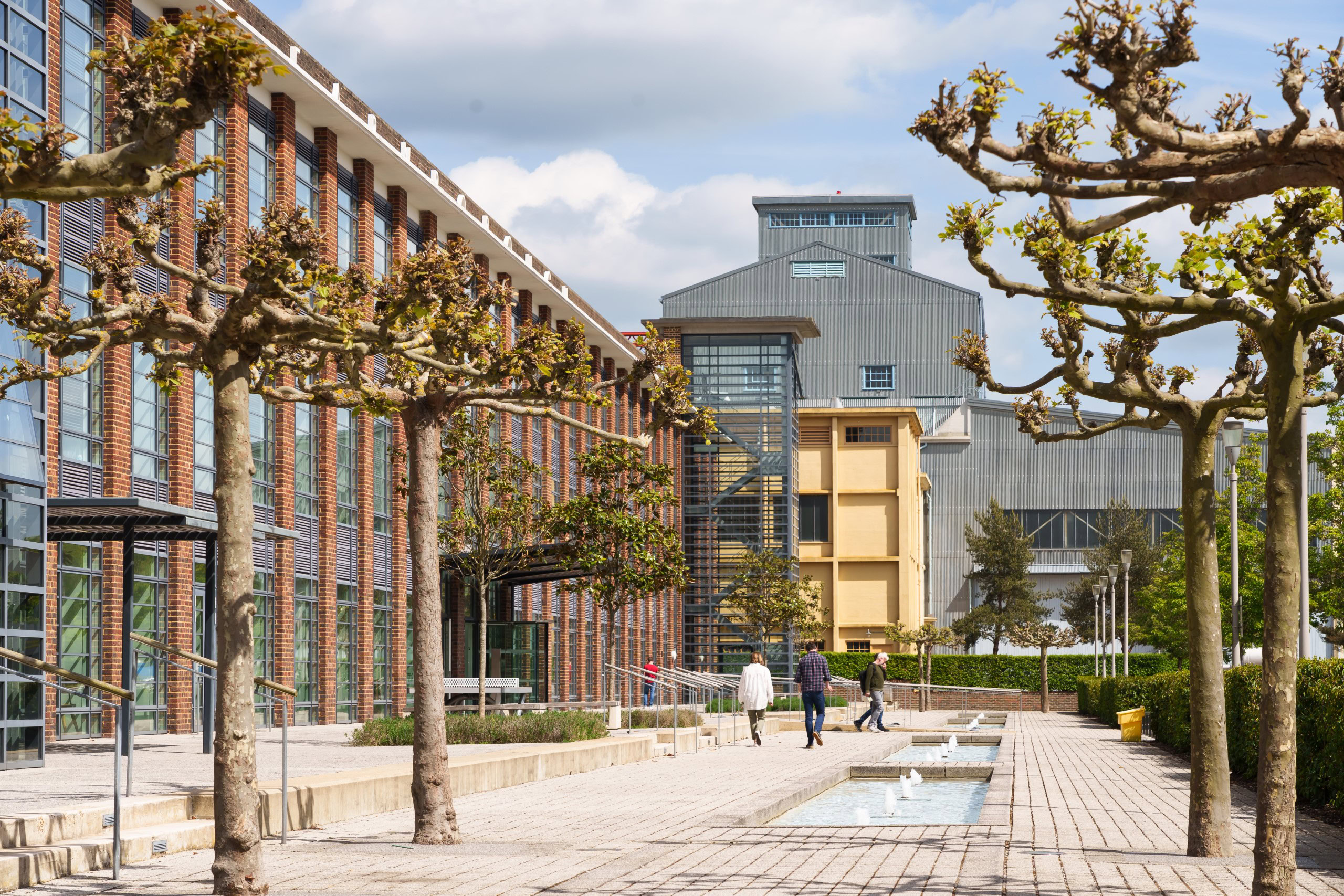 Modern office buildings with brick facades are surrounded by pruned trees and a paved walkway