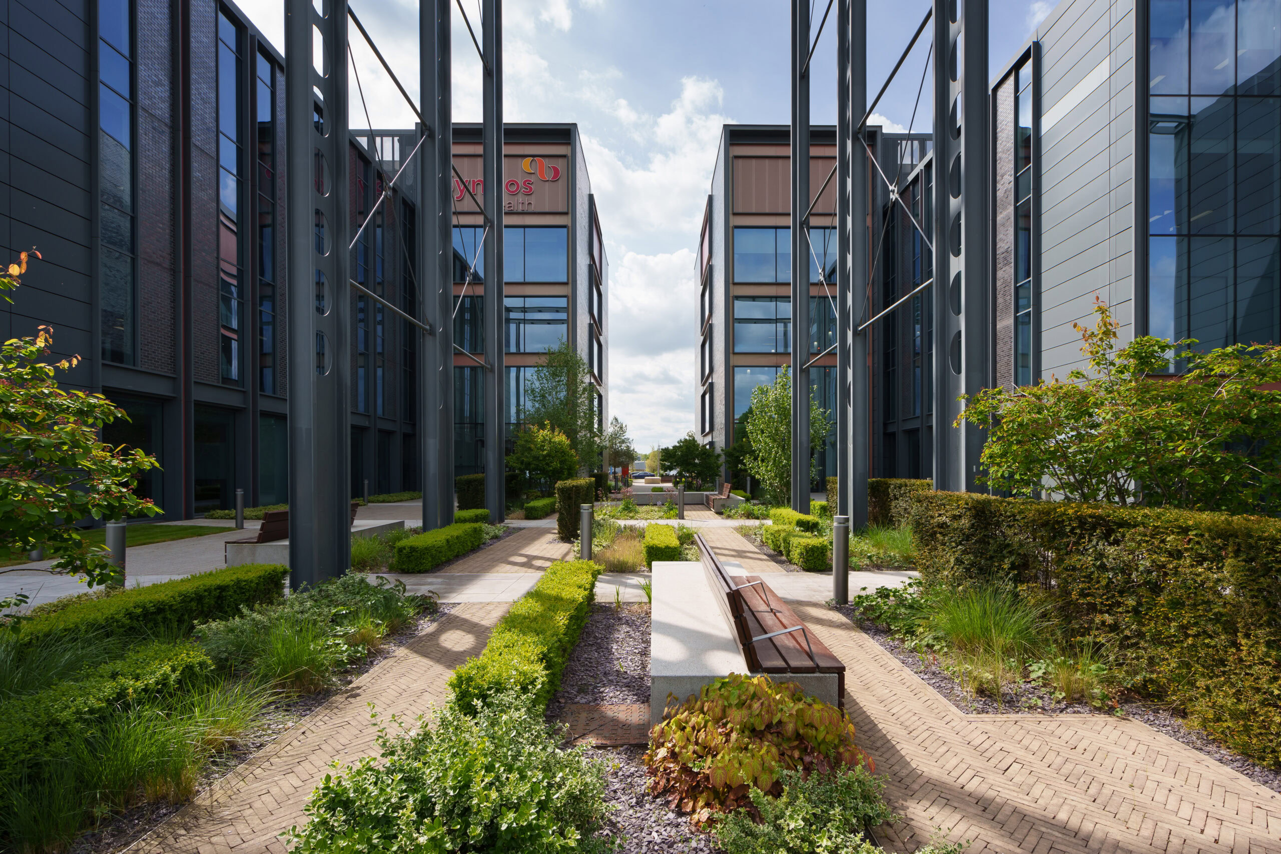 Modern office buildings with glass facades flank a landscaped walkway with benches and greenery.