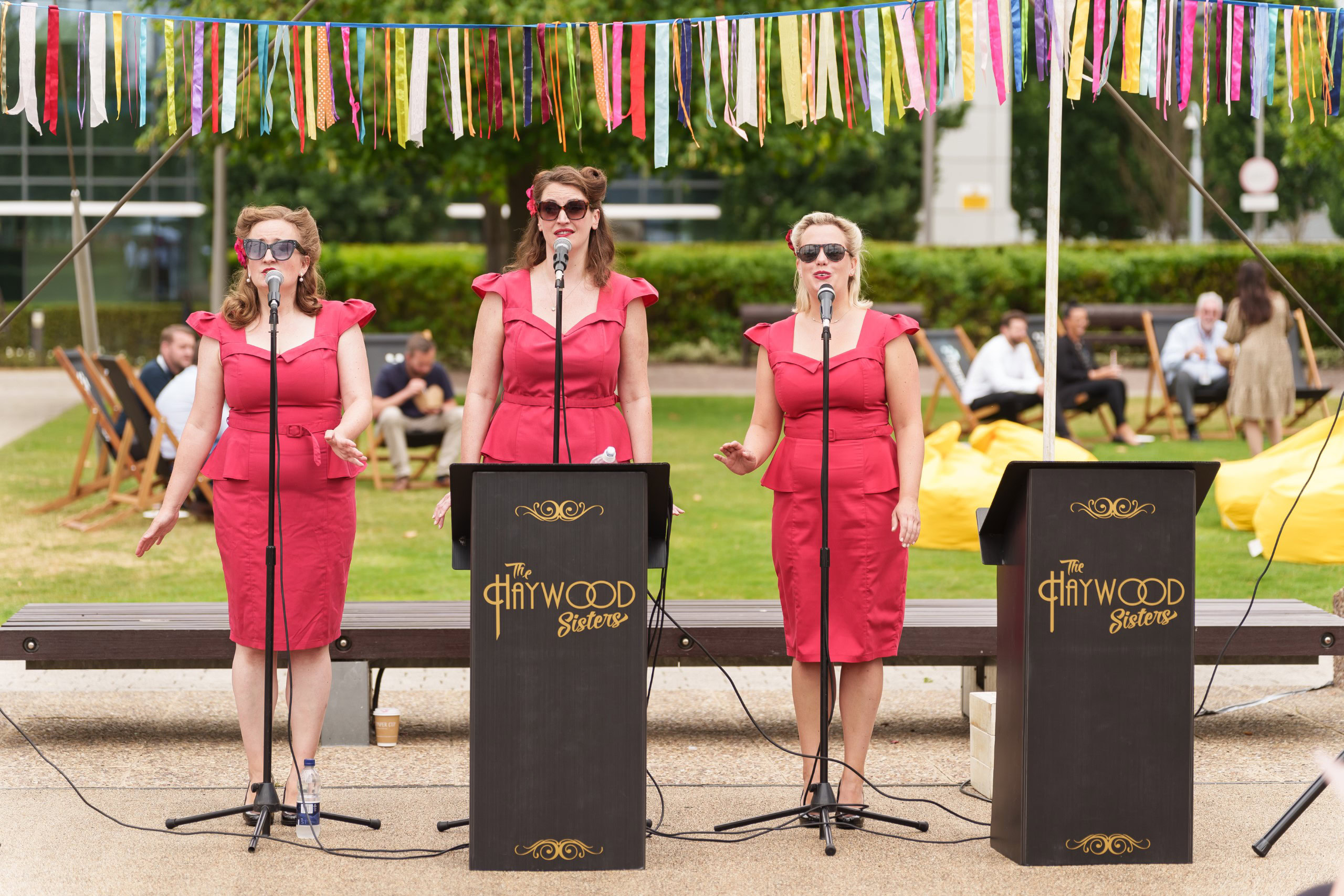 Three women in red dresses sing into microphones on an outdoor stage adorned with colorful ribbons, nestled amidst the backdrop of a bustling business park. People sit in the background, creating a vibrant contrast to the surrounding office buildings.