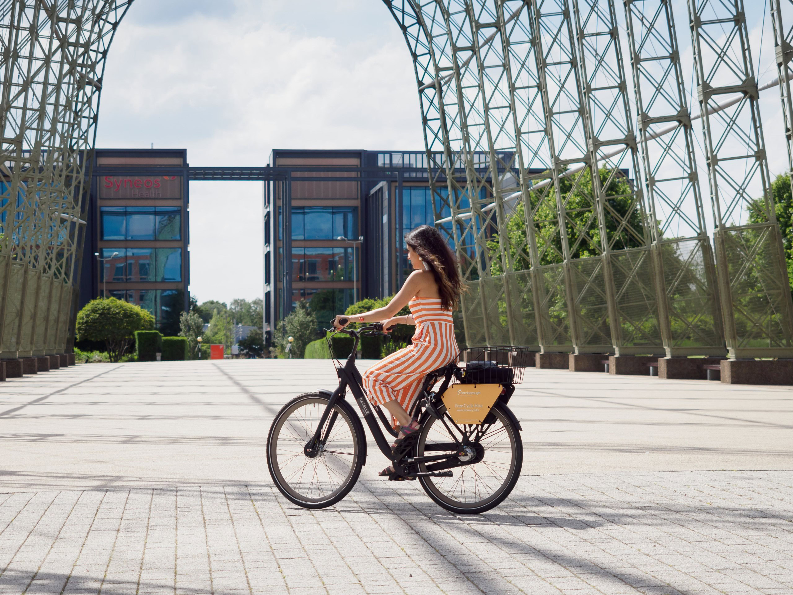 Person riding a black bicycle in front of a large metal structure with buildings in the background.