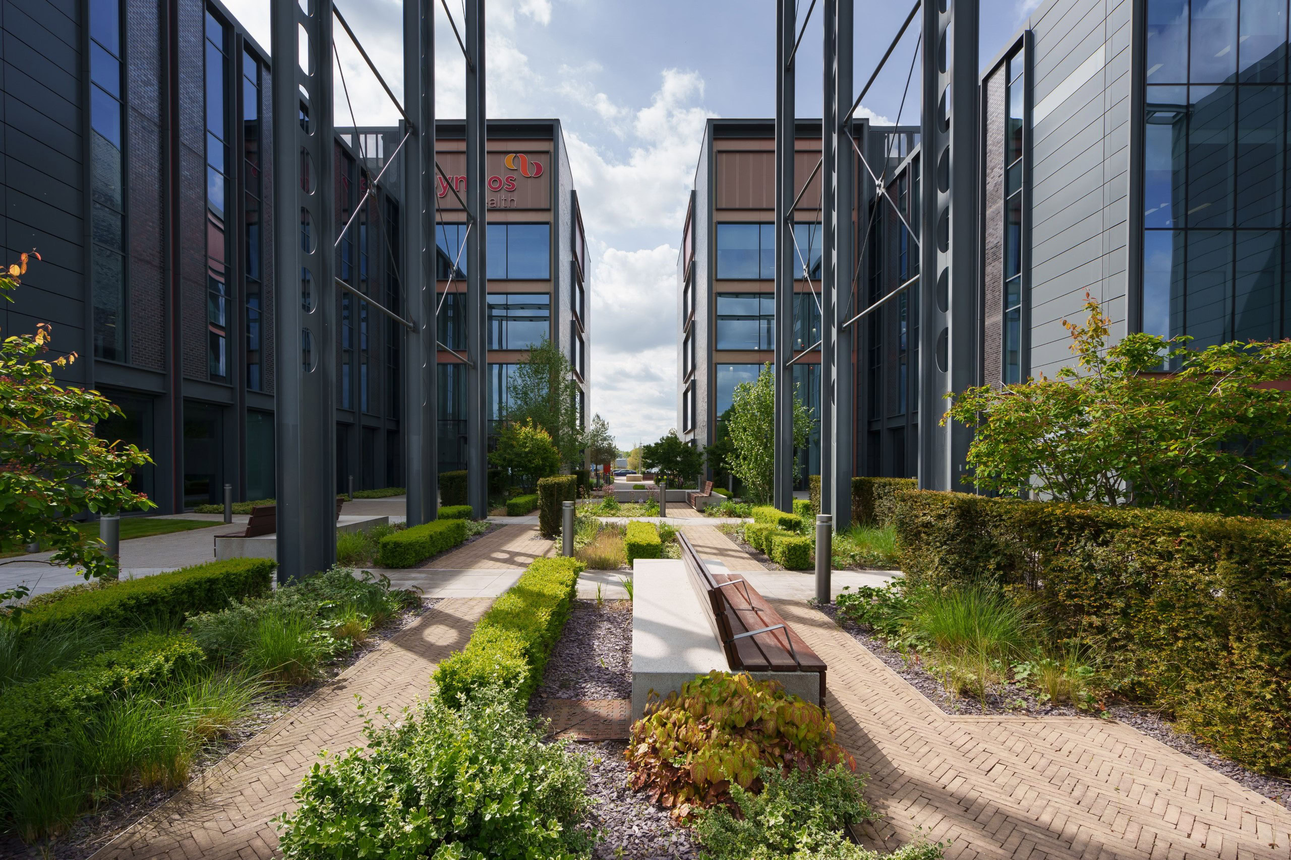 Modern business park courtyard with green hedges and a brick walkway under a partly cloudy sky.