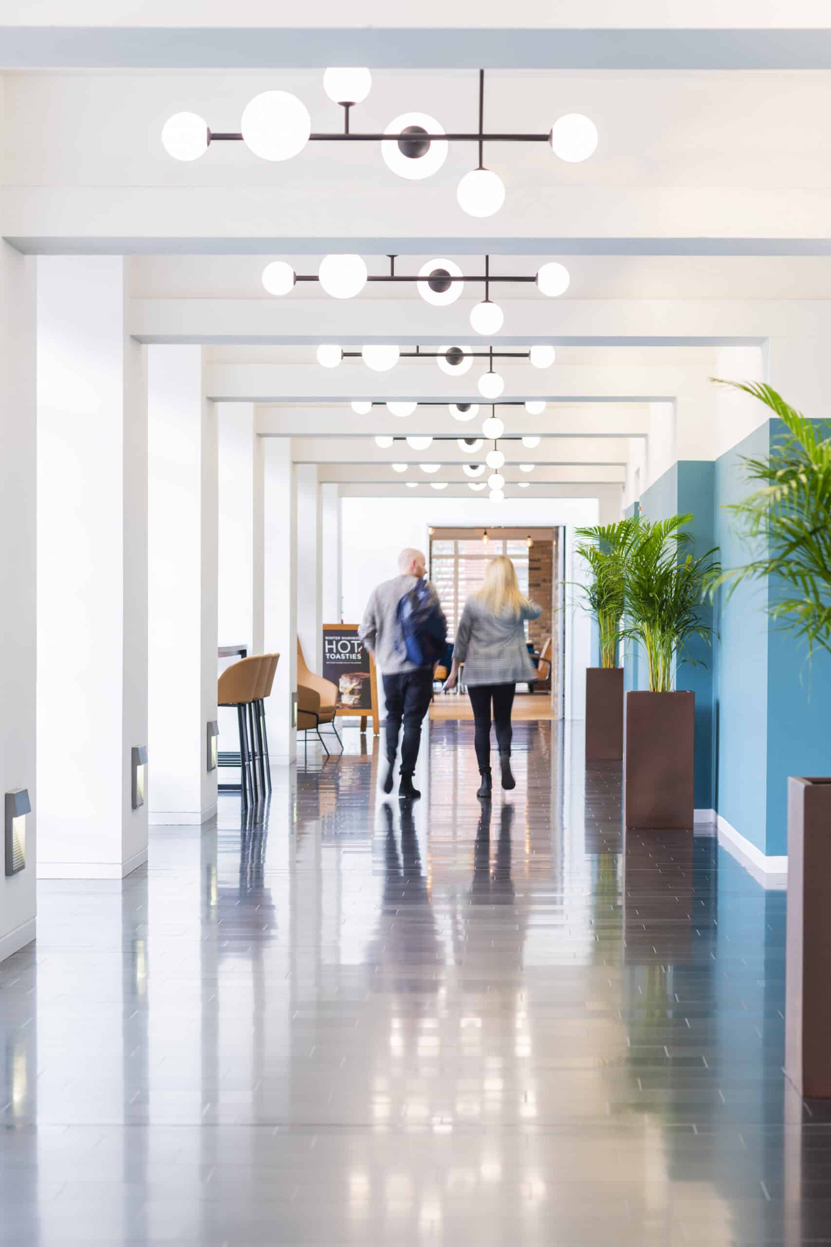 Two people walk down a modern hallway featuring sleek flooring, potted plants, and light fixtures