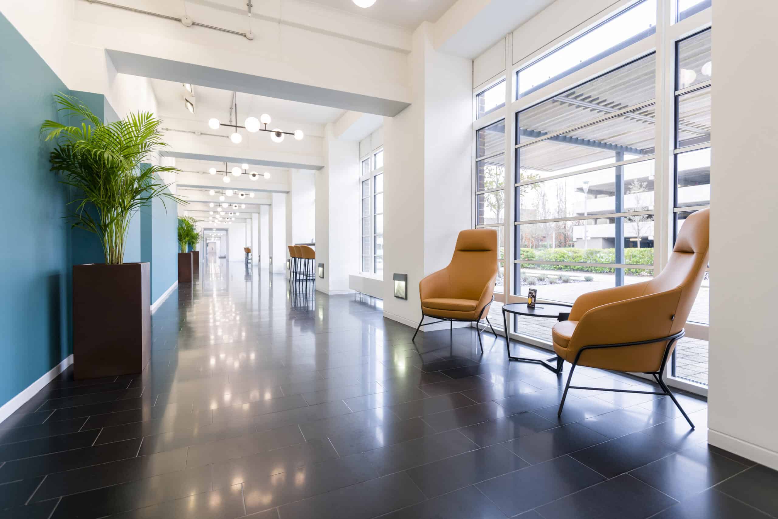 Modern corridor with large windows, black tiled floor, 2 brown chairs, and small tables with plants