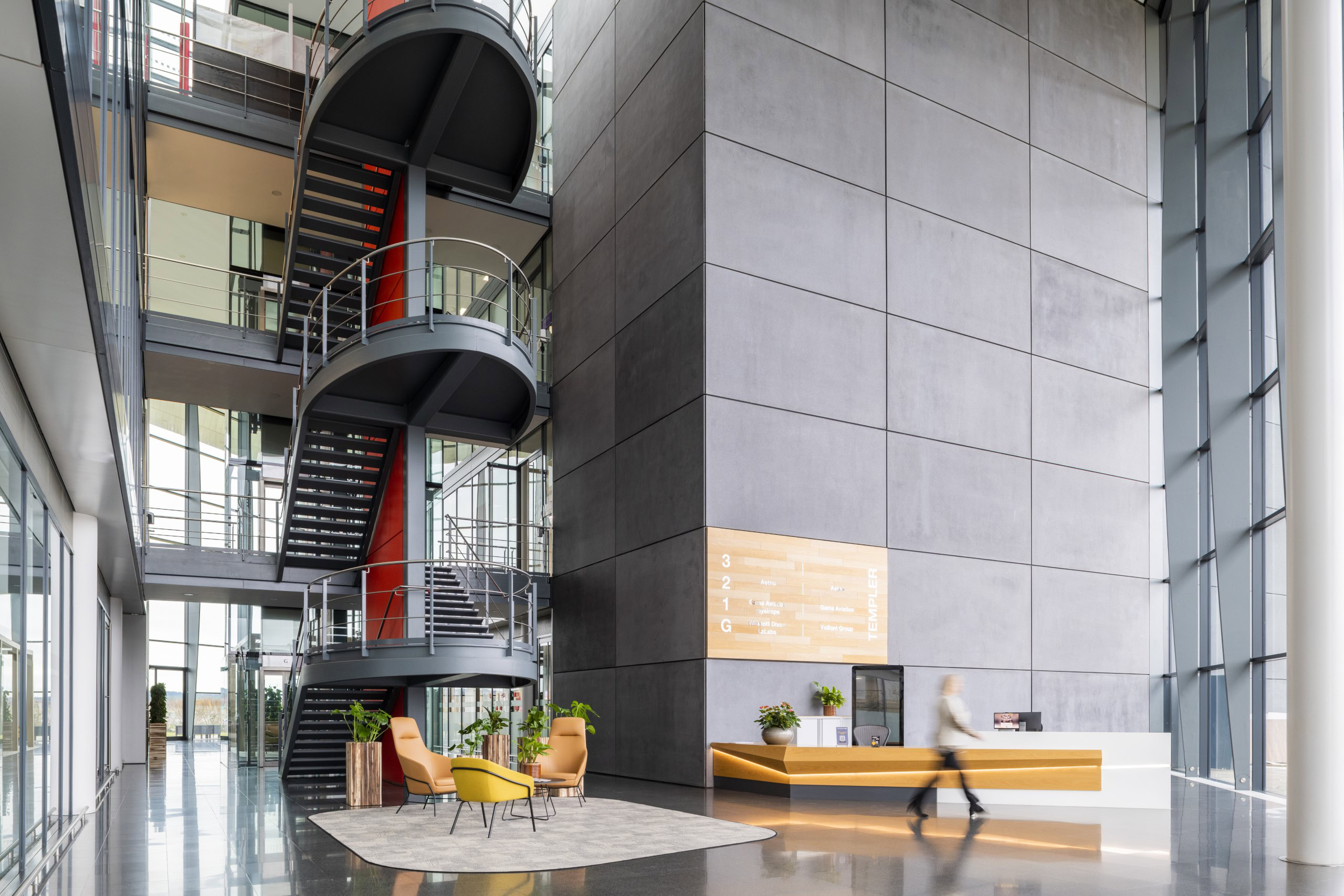 A modern office building lobby with a tall gray wall, spiral staircase, and large windows