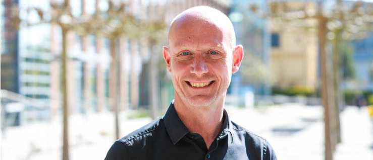 A man with a shaved head smiles outdoors, wearing a dark shirt
