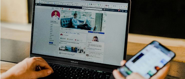 A person in an office building uses a laptop with Facebook on the screen while holding a smartphone