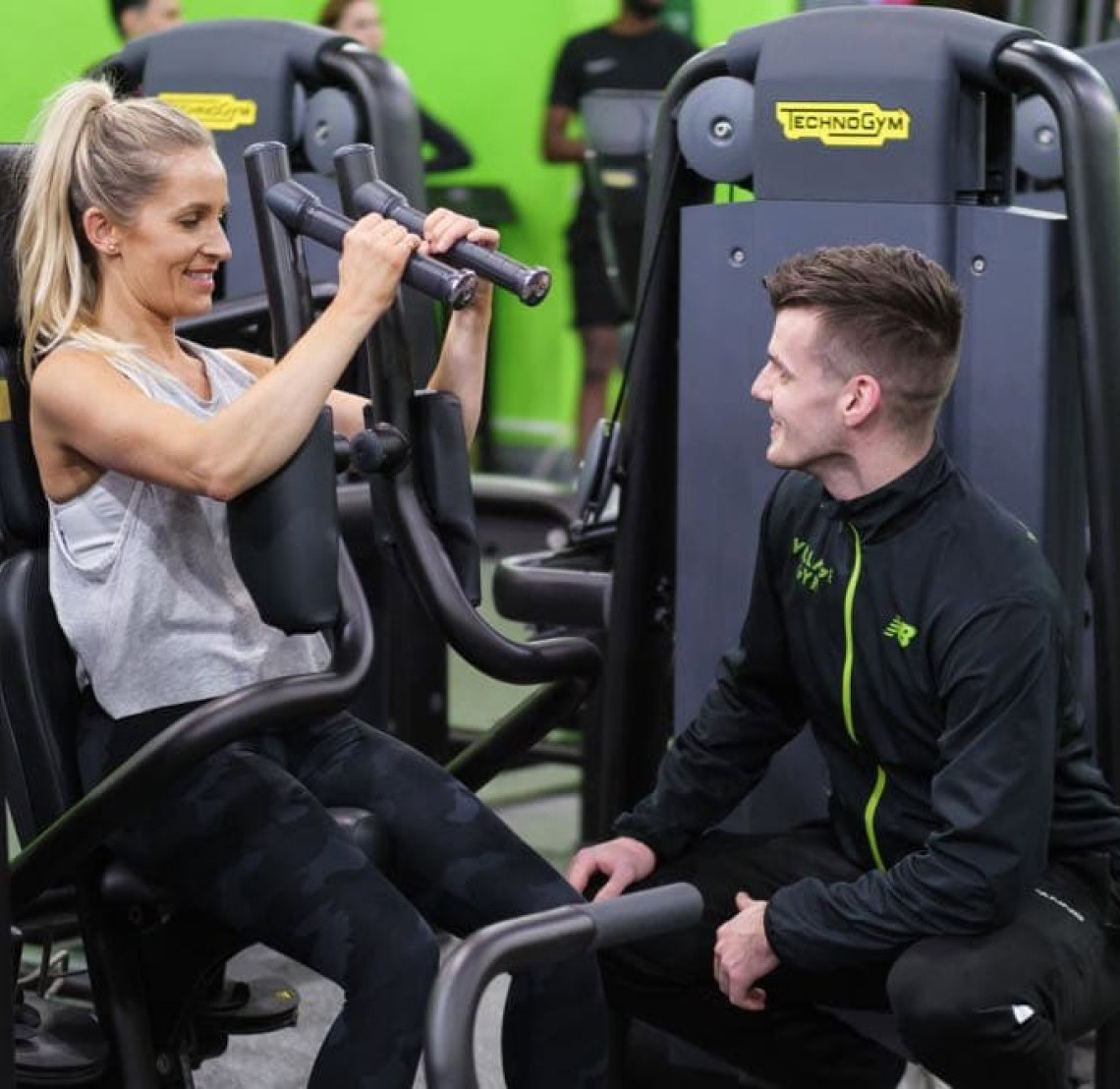 A woman uses an exercise machine at the gym, while a trainer kneels beside her, providing guidance