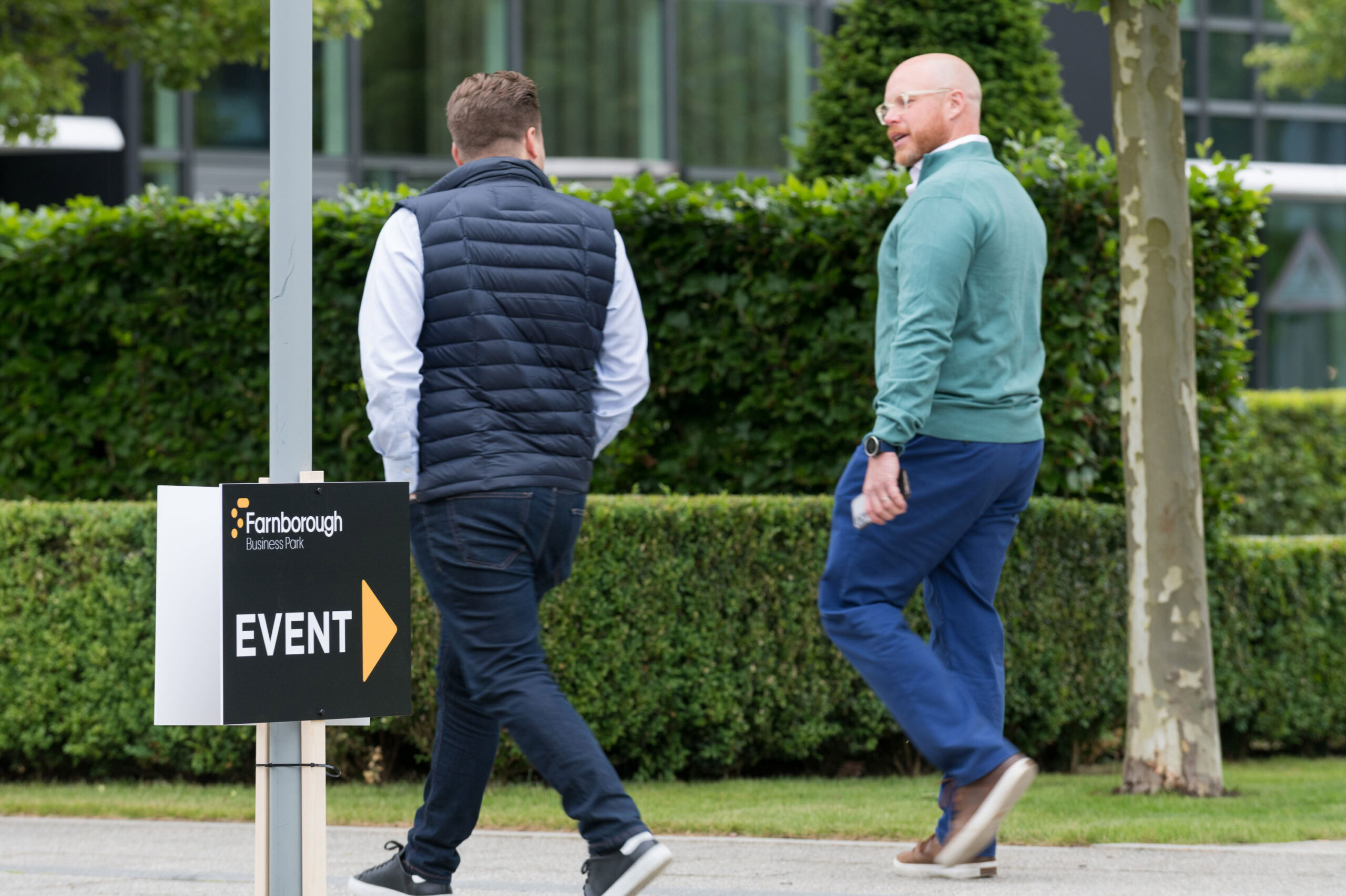 Two men walk past a sign at Farnborough Business Park, surrounded by modern office buildings, with "Event" and an arrow pointing right. One wears a navy vest, the other a teal sweater. Bushes and trees are in the background.