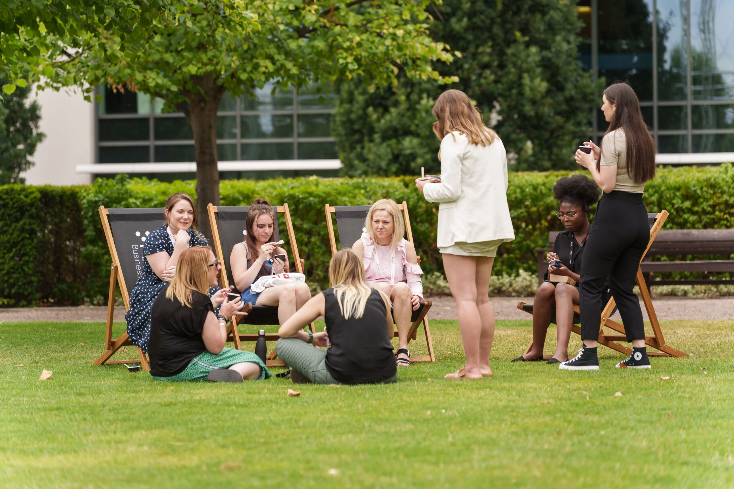 A group of people enjoying a sunny afternoon on the lawn of a business park, with trees in the background. Some are seated on deck chairs, while others stand or sit on the grass, holding drinks, taking a break from their nearby office building.