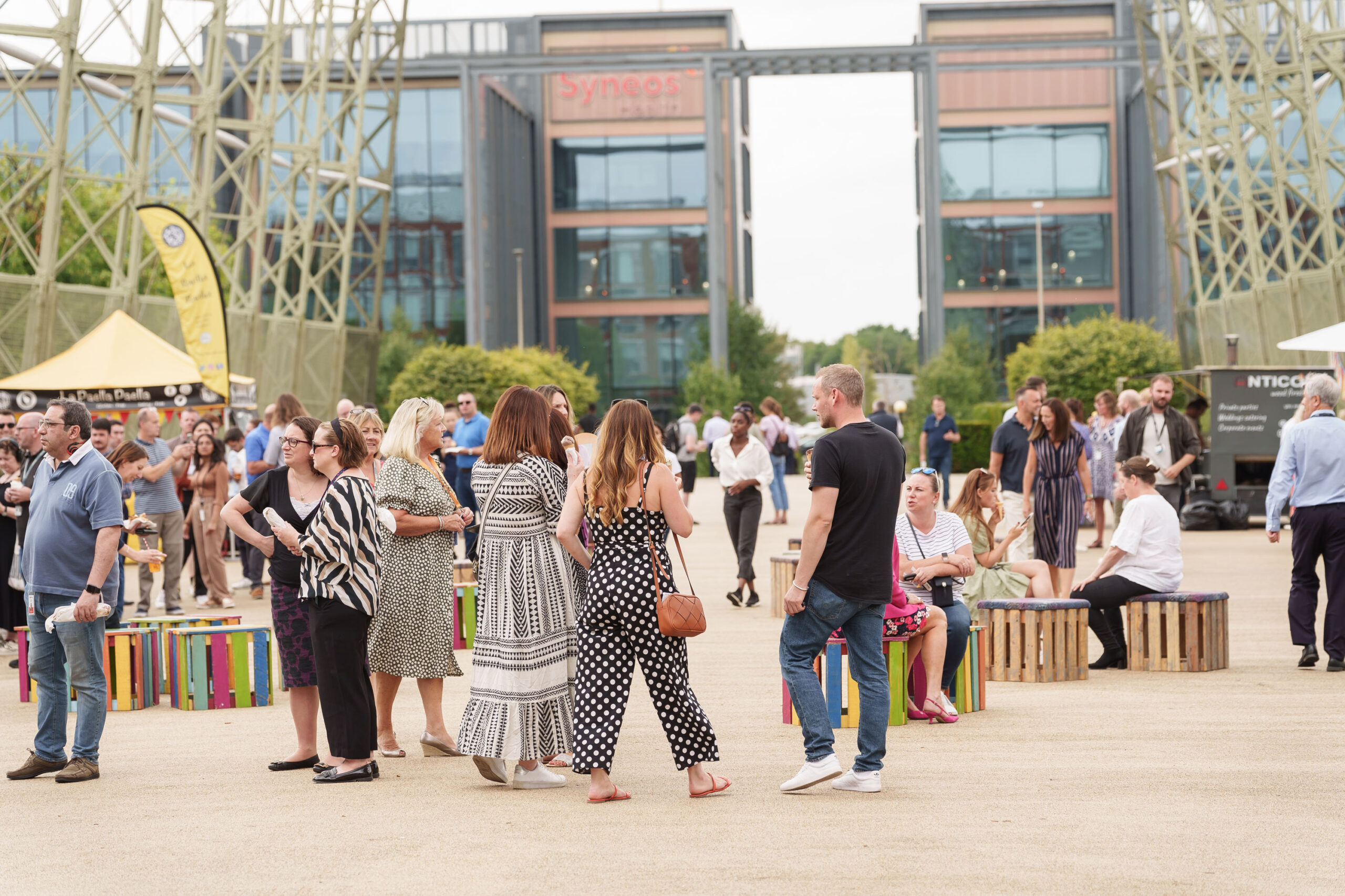 People gathered in an outdoor area of a bustling business park, modern office buildings in the background, some sitting on wooden benches while others stand and chat.
