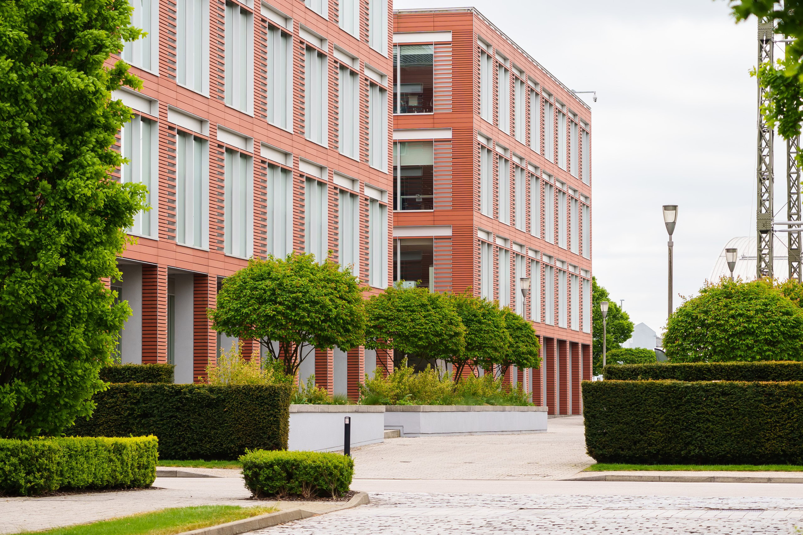 A modern, red-brick office building with large windows surrounded by neatly trimmed trees and hedges