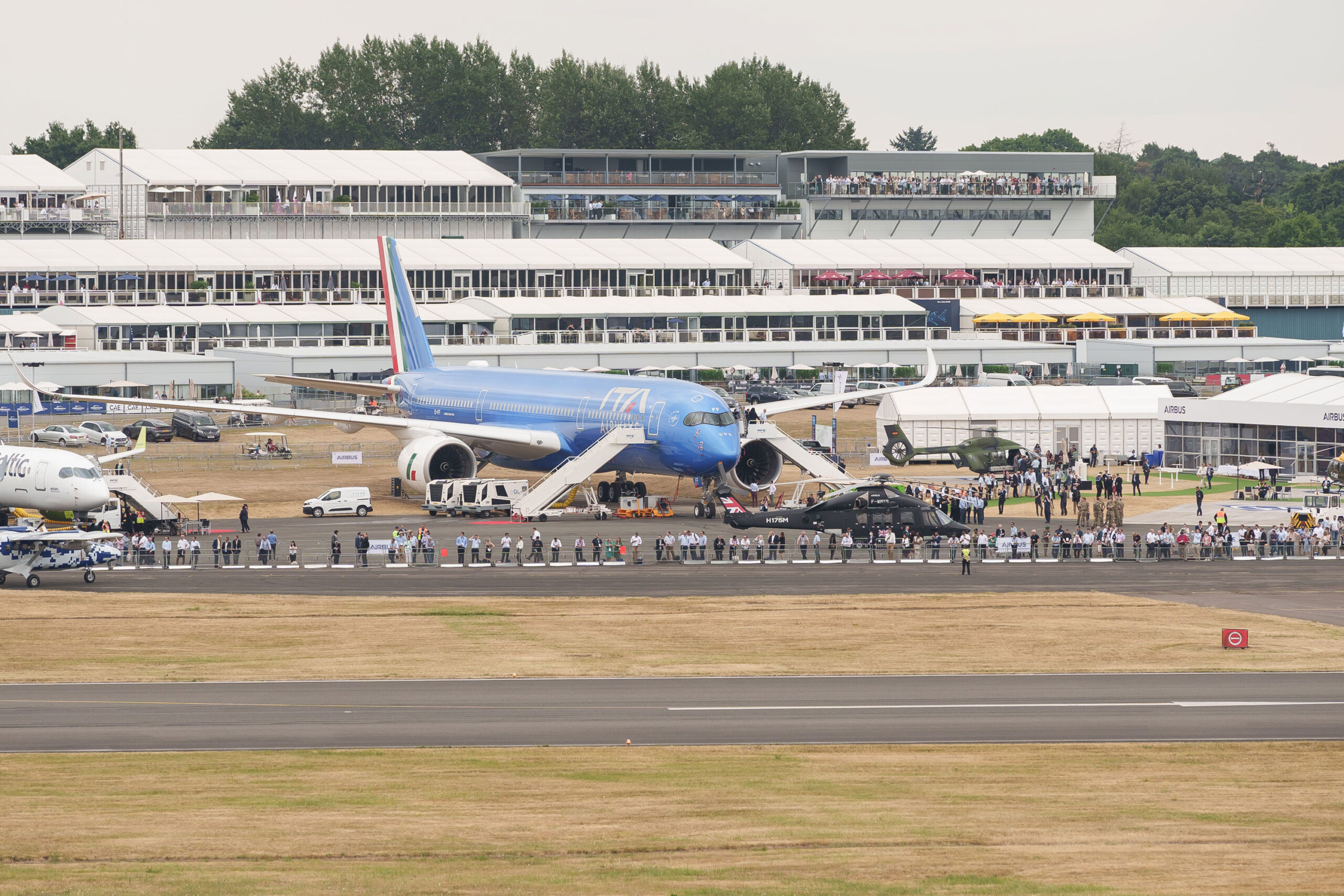 A blue aircraft is on display at an airshow, attracting a crowd. In the background, several white tents and a grandstand rise like an office building in a business park.