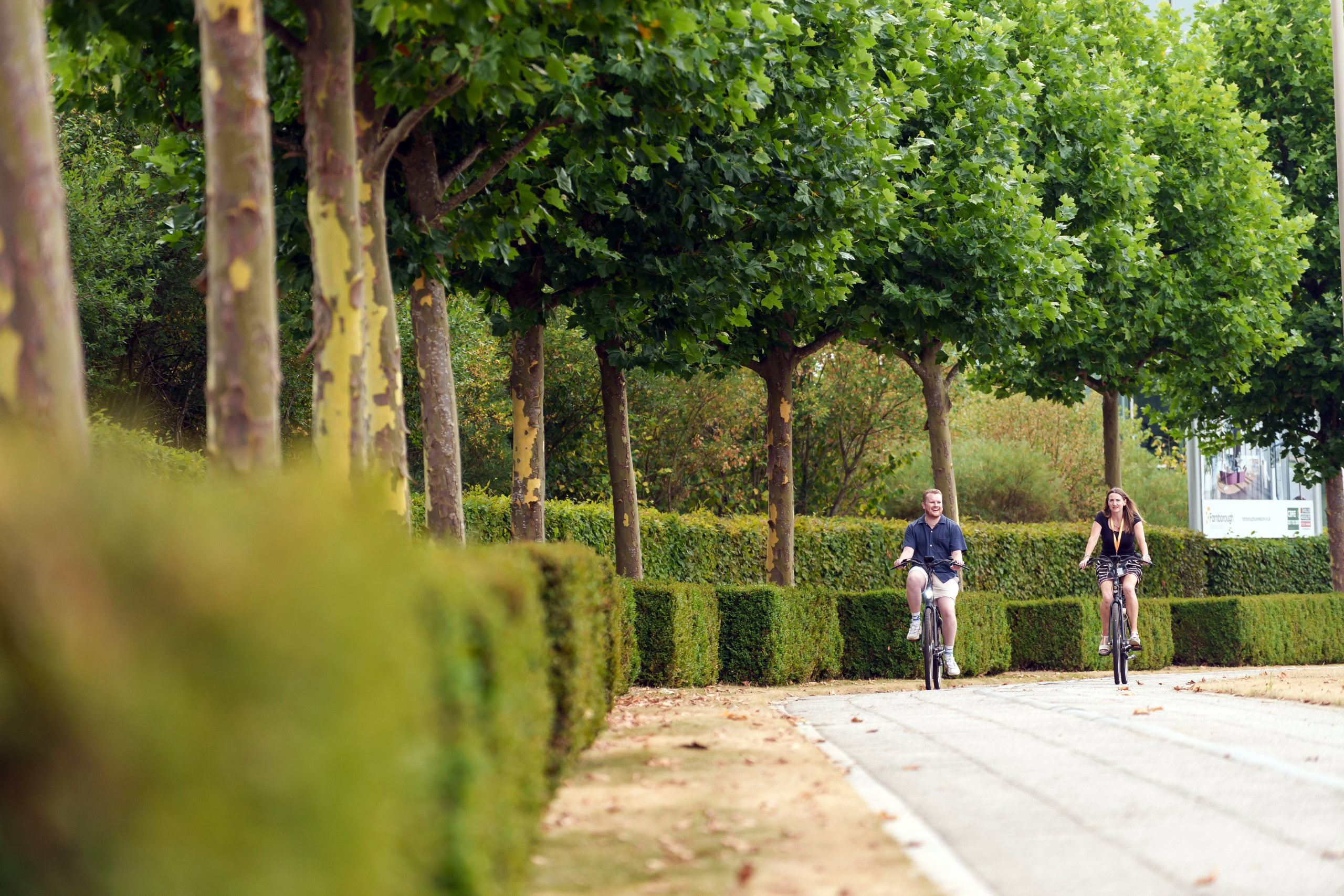 Two people ride bicycles on a tree-lined path near the edge of the business park.
