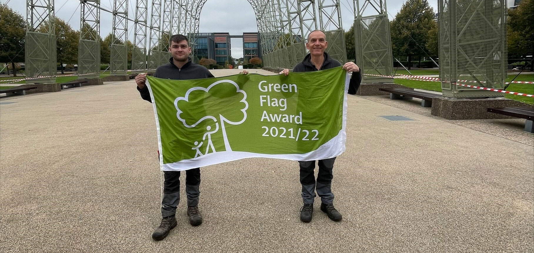 Two people hold a "Green Flag Award 2021/22" banner outdoors with office buildings in the background
