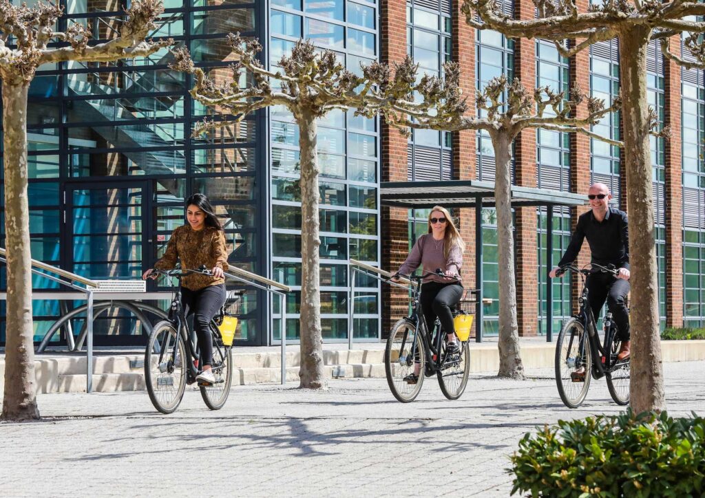 Three people ride bikes on a paved path near a modern glass office building