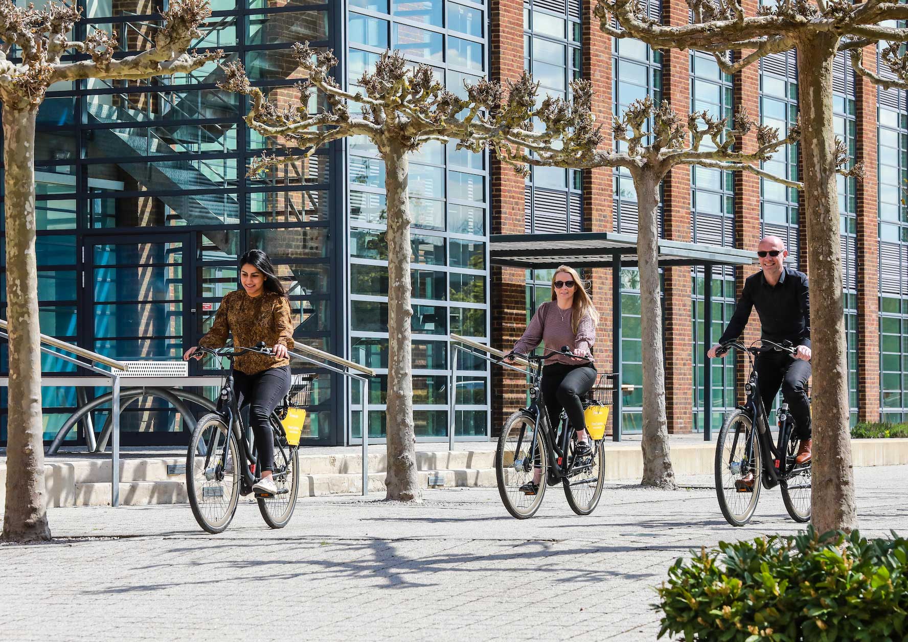 Three people ride bikes on a paved path near a modern glass office building