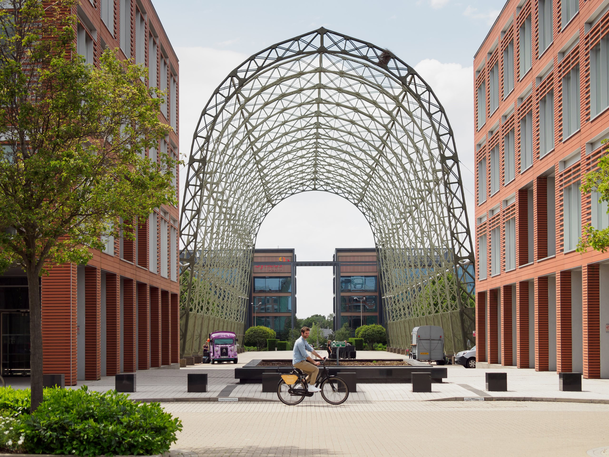 A person rides a bicycle in front of a large arched structure between two red brick office buildings