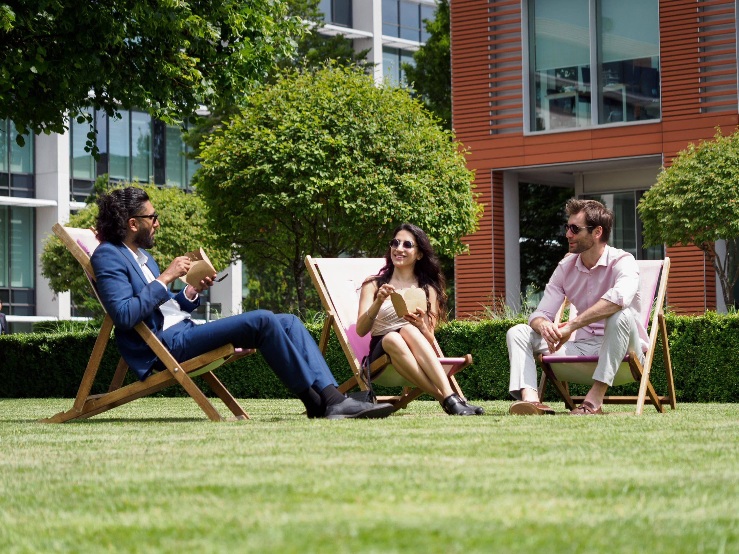 Three people sit on deck chairs in a business park's grassy area, engaging in conversation