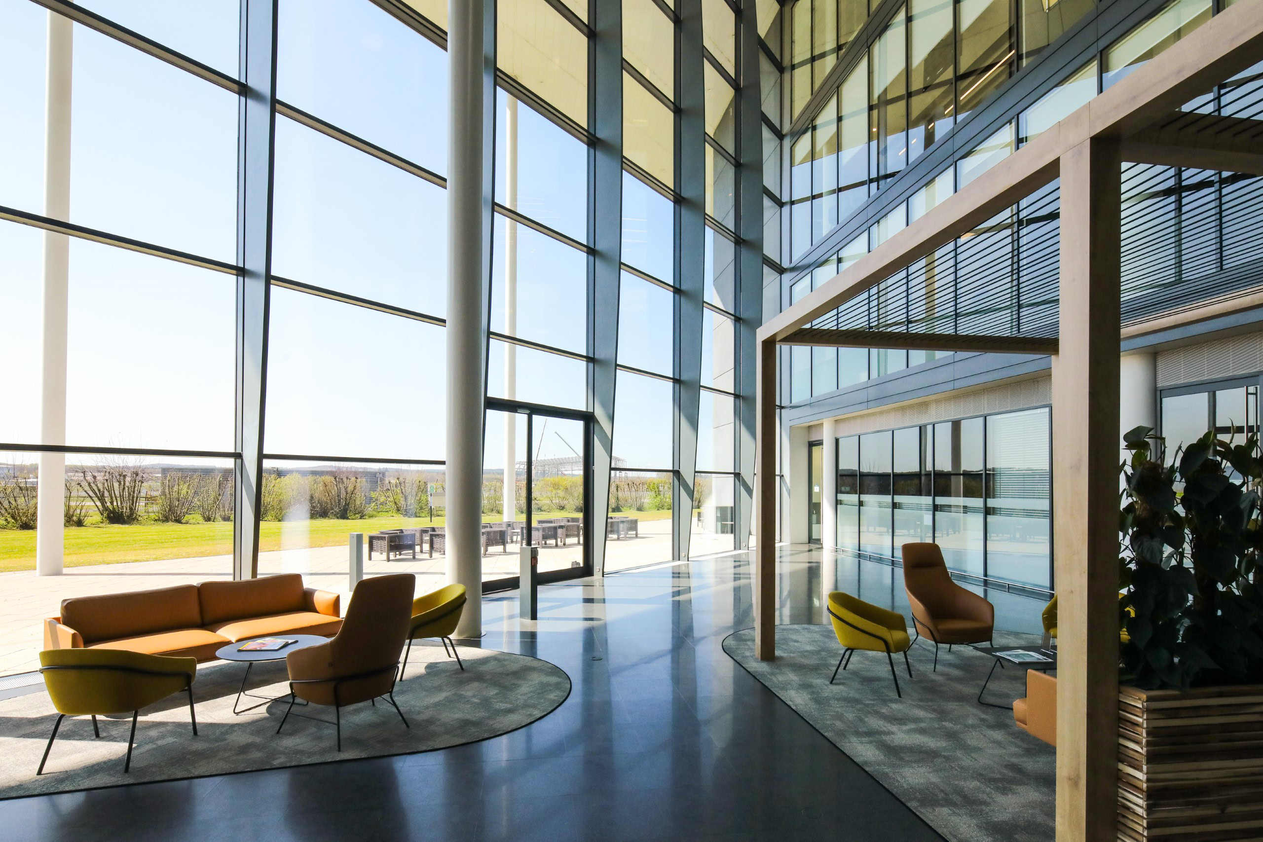 In a modern office building lobby, large glass windows illuminate yellow chairs and seating areas