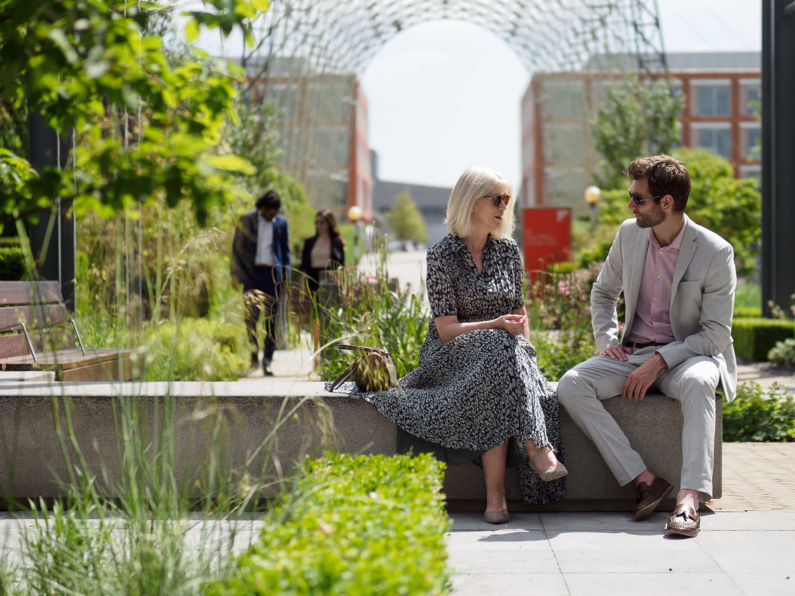 Two people sit on a stone bench in a garden setting within a business park, engaged in conversation