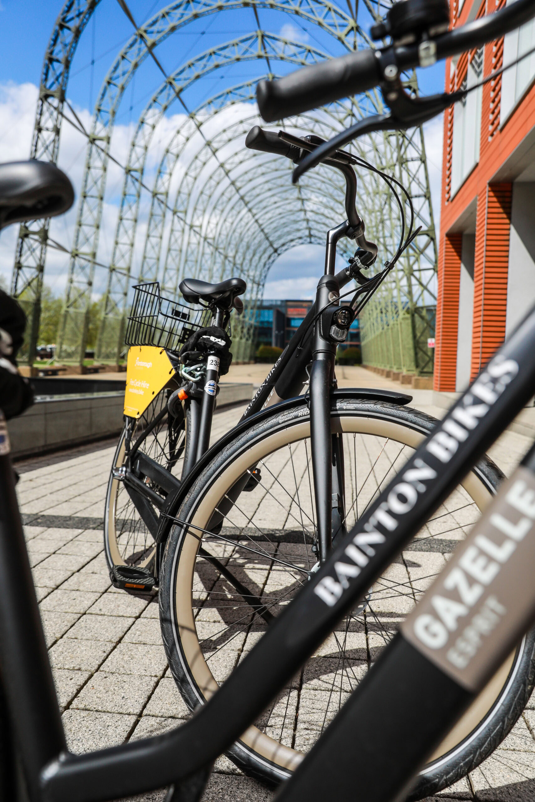 Black bicycles parked on a paved path in front of a metal arch structure and a red office building
