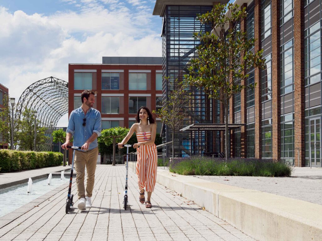 A man and a woman walk alongside scooters on a paved path next to an office building