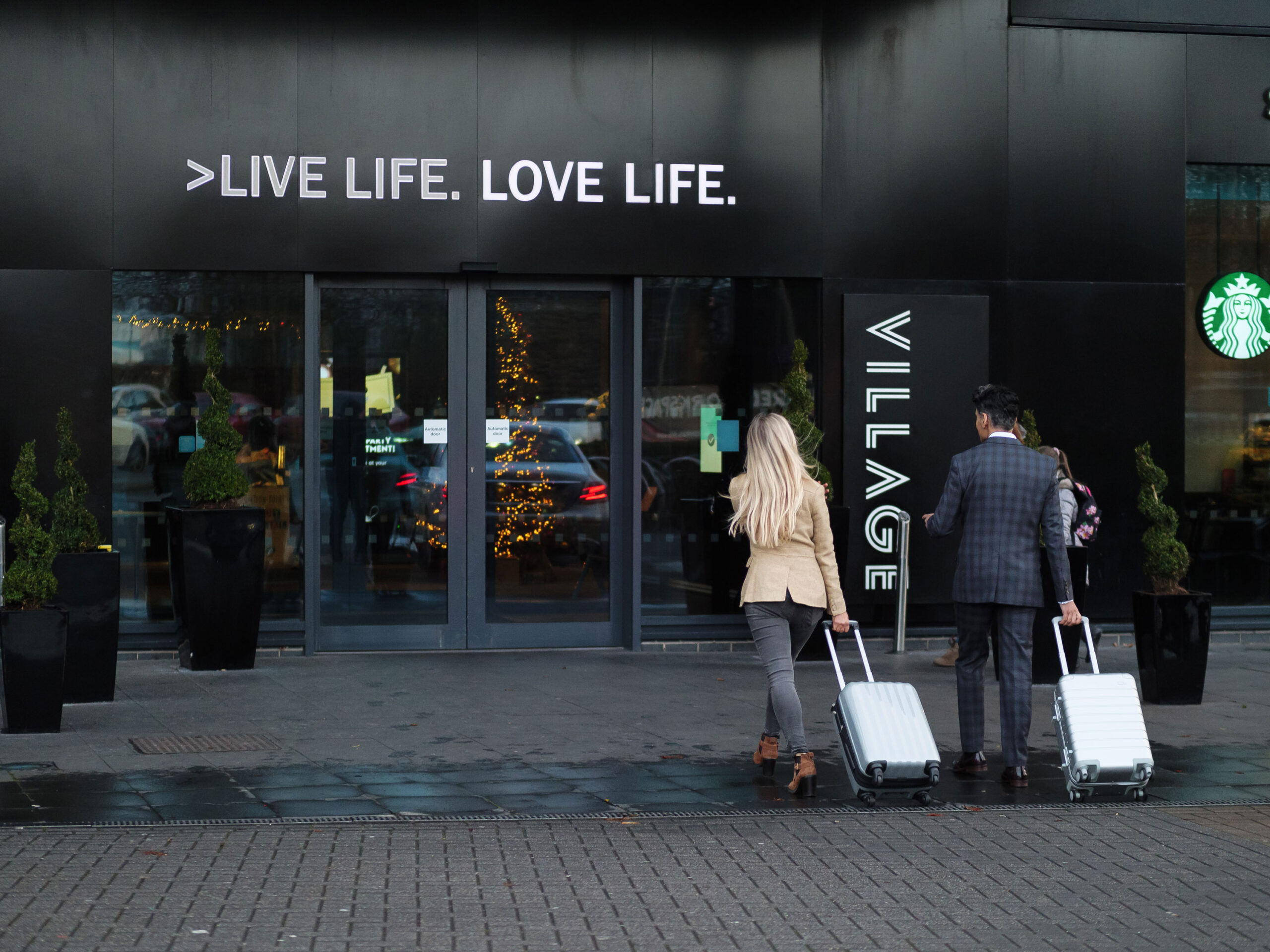Two people with suitcases walk towards a hotel entrance labelled Village"
