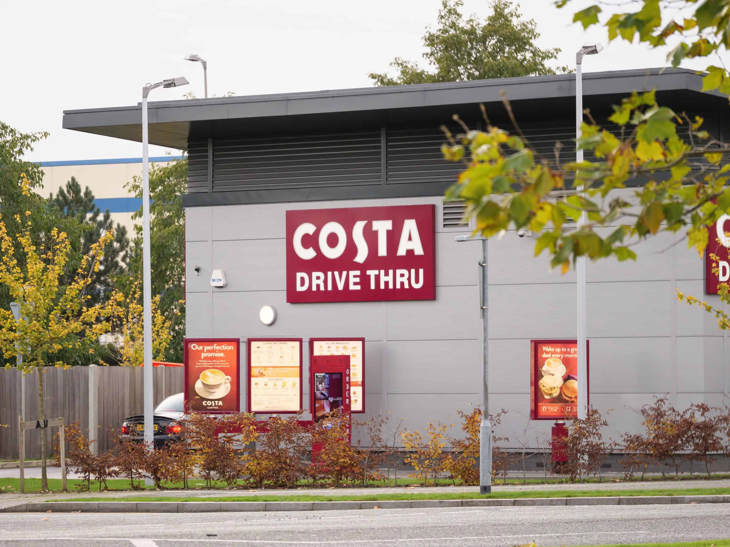 Costa Drive Thru building exterior with signage and a menu board, surrounded by trees & parked cars