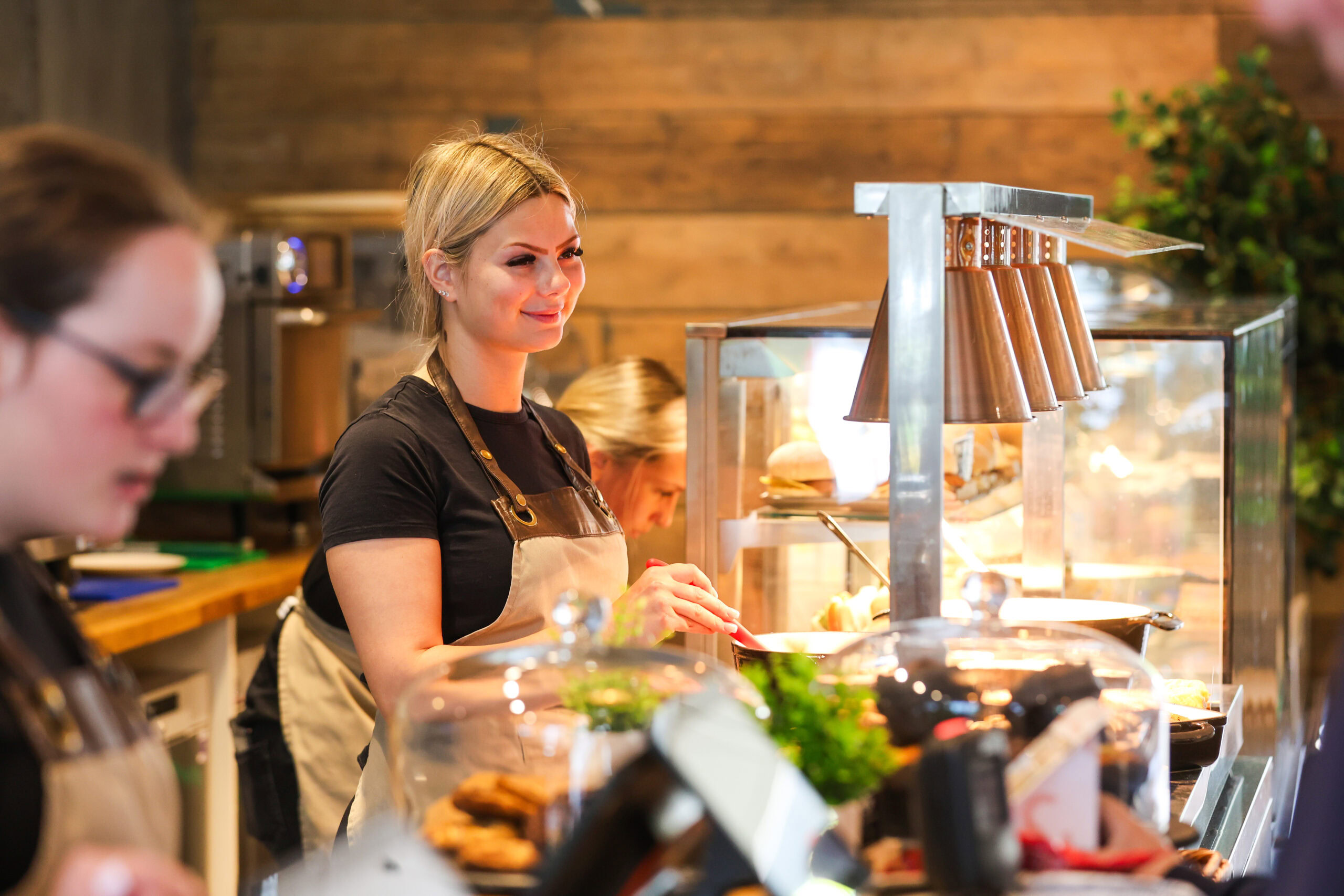 A woman in an apron serves food behind a counter in a brightly lit café or restaurant.