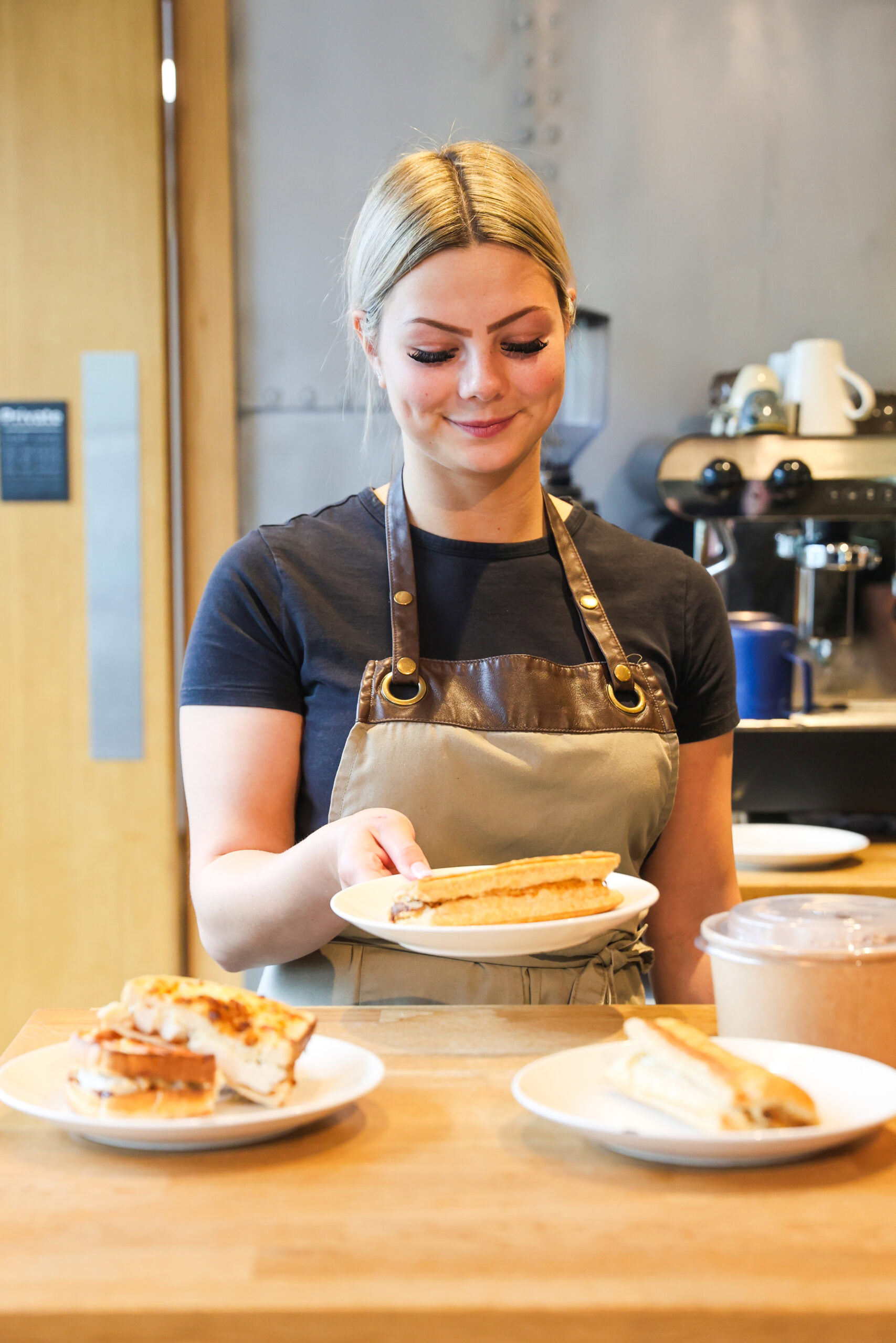 A woman in an apron serves a toasted sandwich on a plate in a café kitchen.