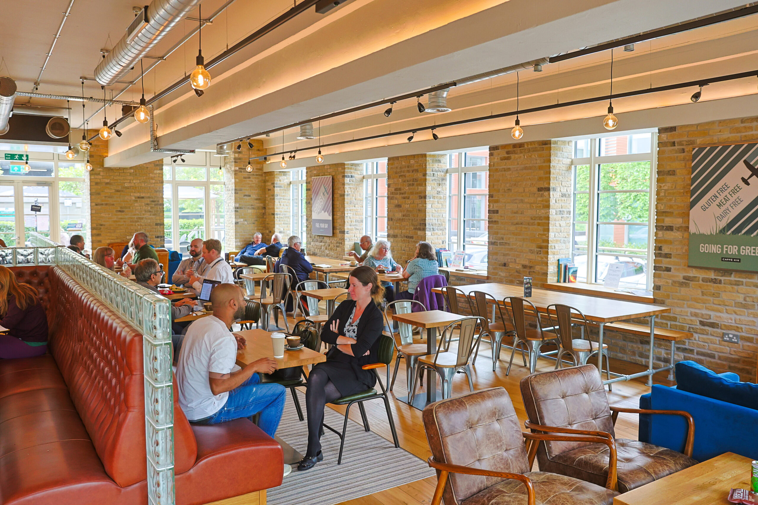 People sit and talk at tables in a bright, modern café with large windows and exposed brick walls.