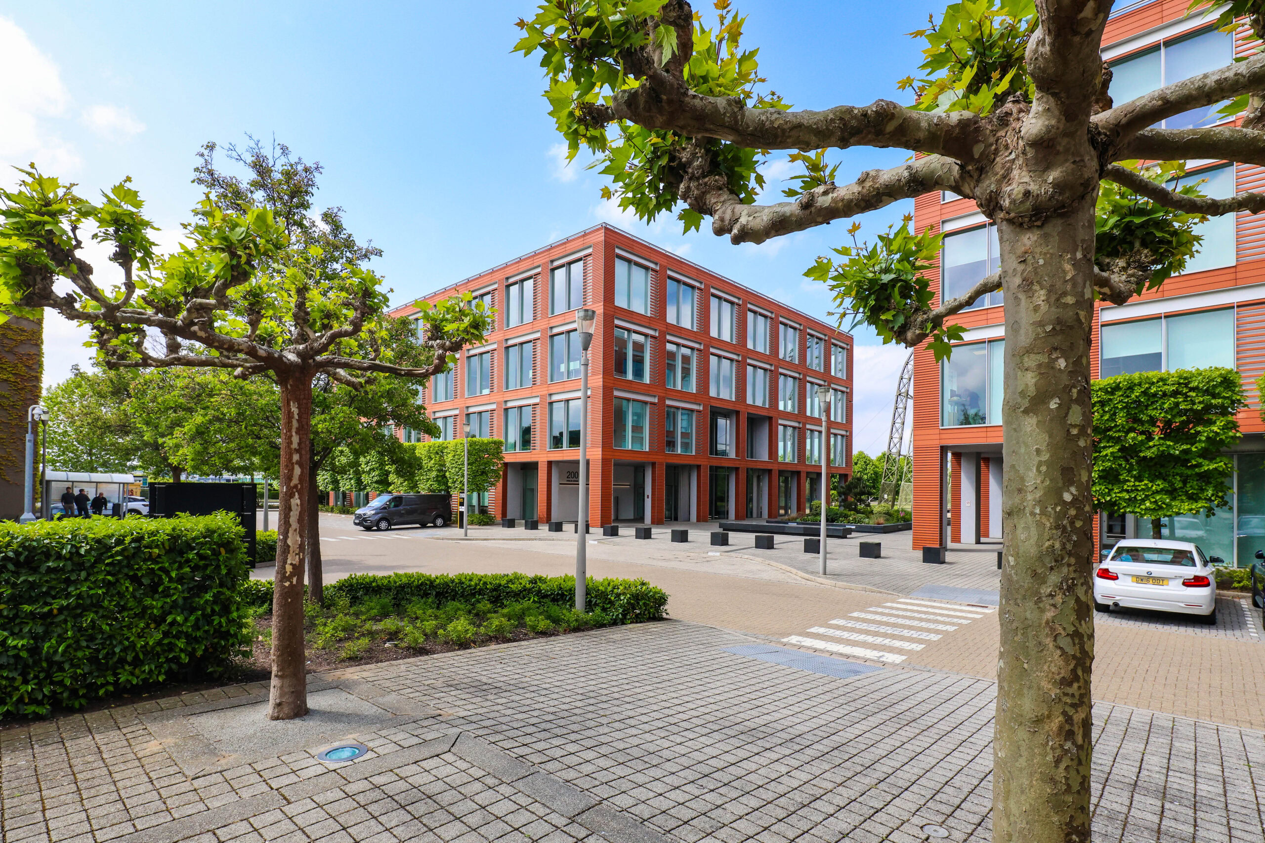 Modern brick office building with large windows, surrounded by trees and parked cars on a sunny day.