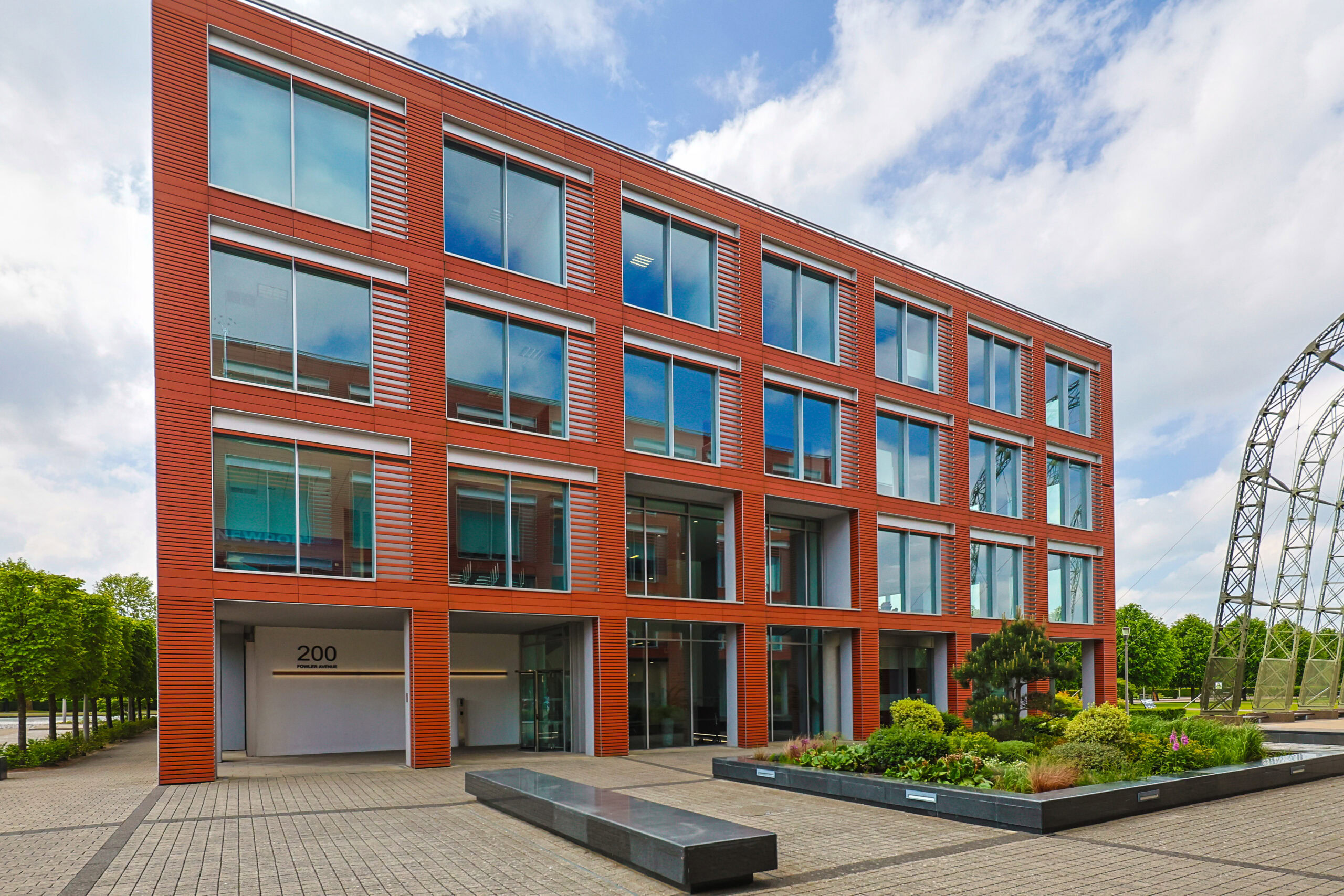 Modern red office building with large windows, landscaped entrance, and cloudy sky background.