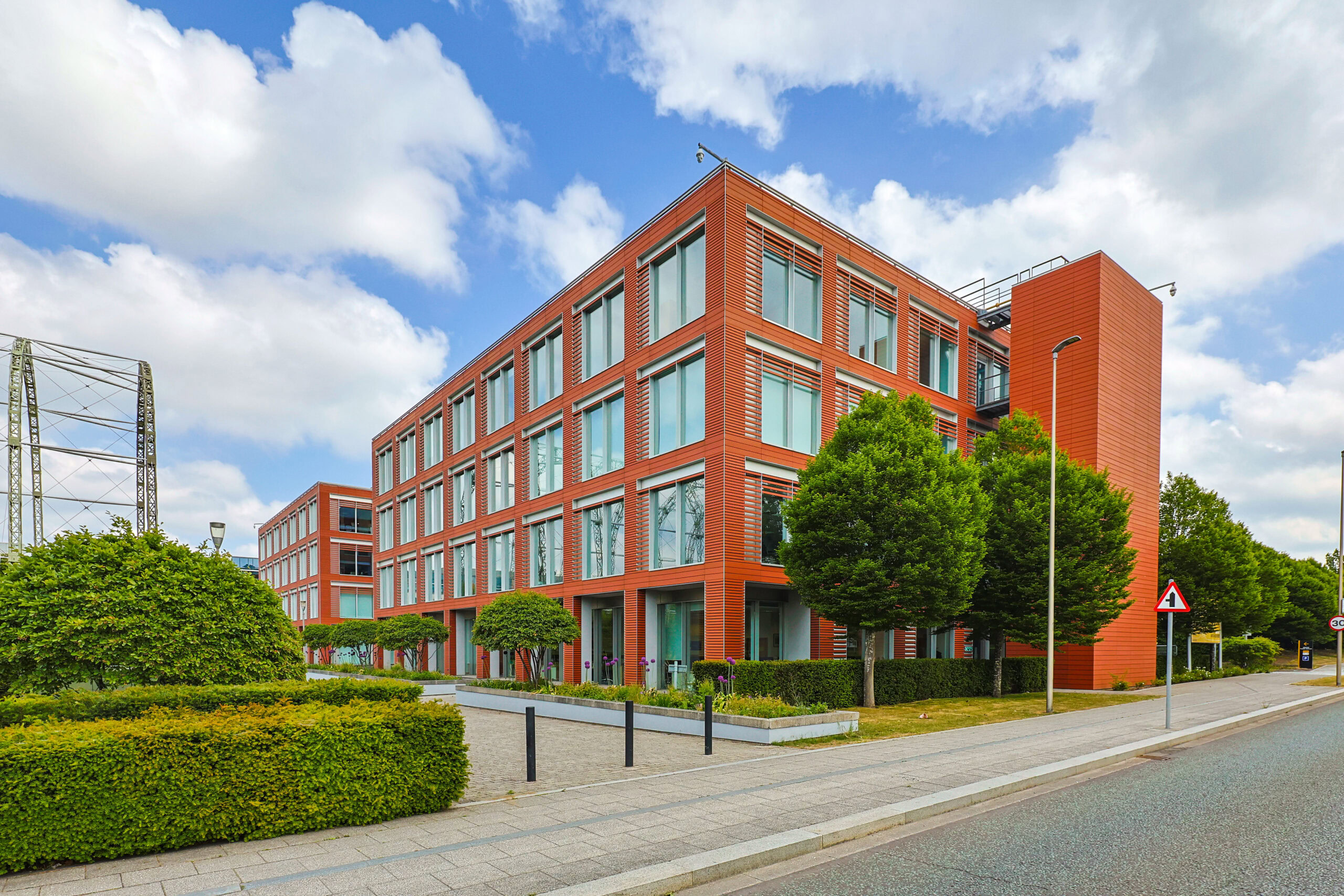 Modern red-brick office building with large windows, trees, and sidewalk under a partly cloudy sky.