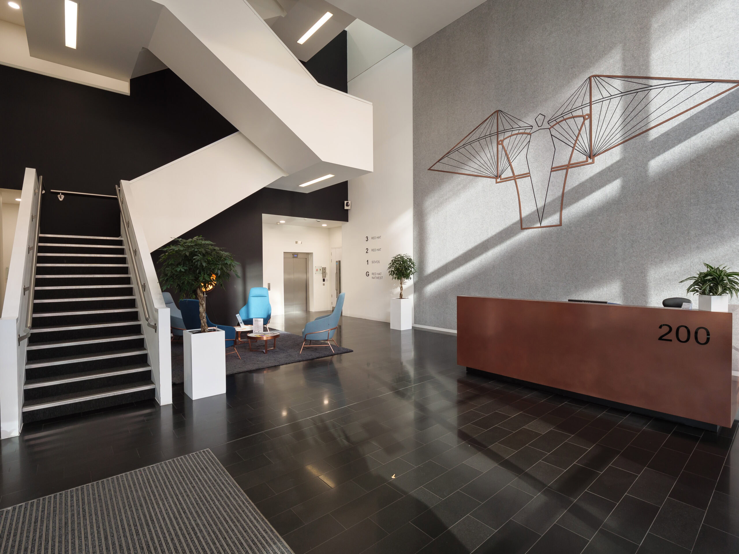 Modern office lobby with staircase, reception desk labeled "200," chairs, and geometric wall art.