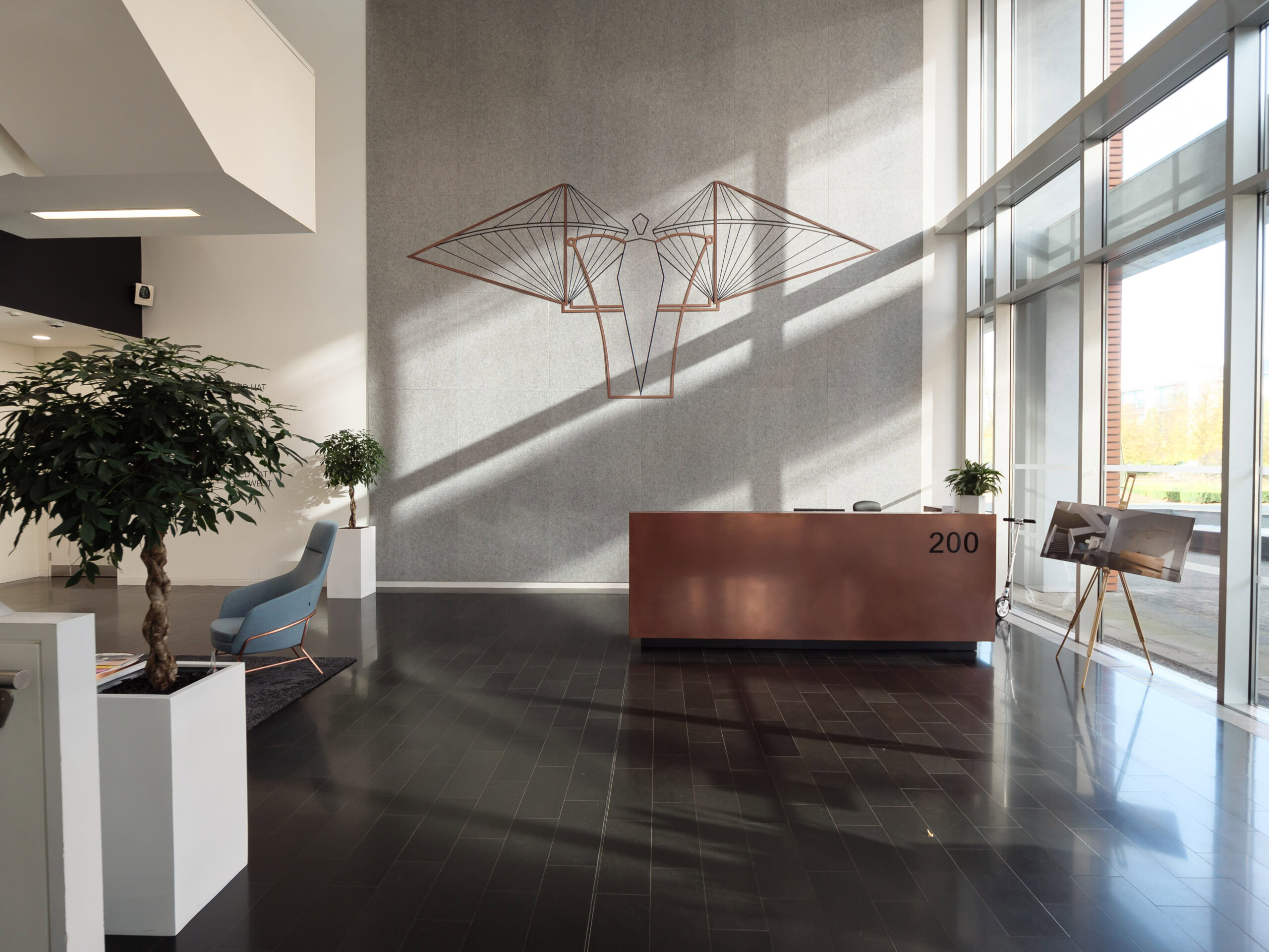 Modern office lobby with a copper reception desk, wall art, large windows, and potted plants.