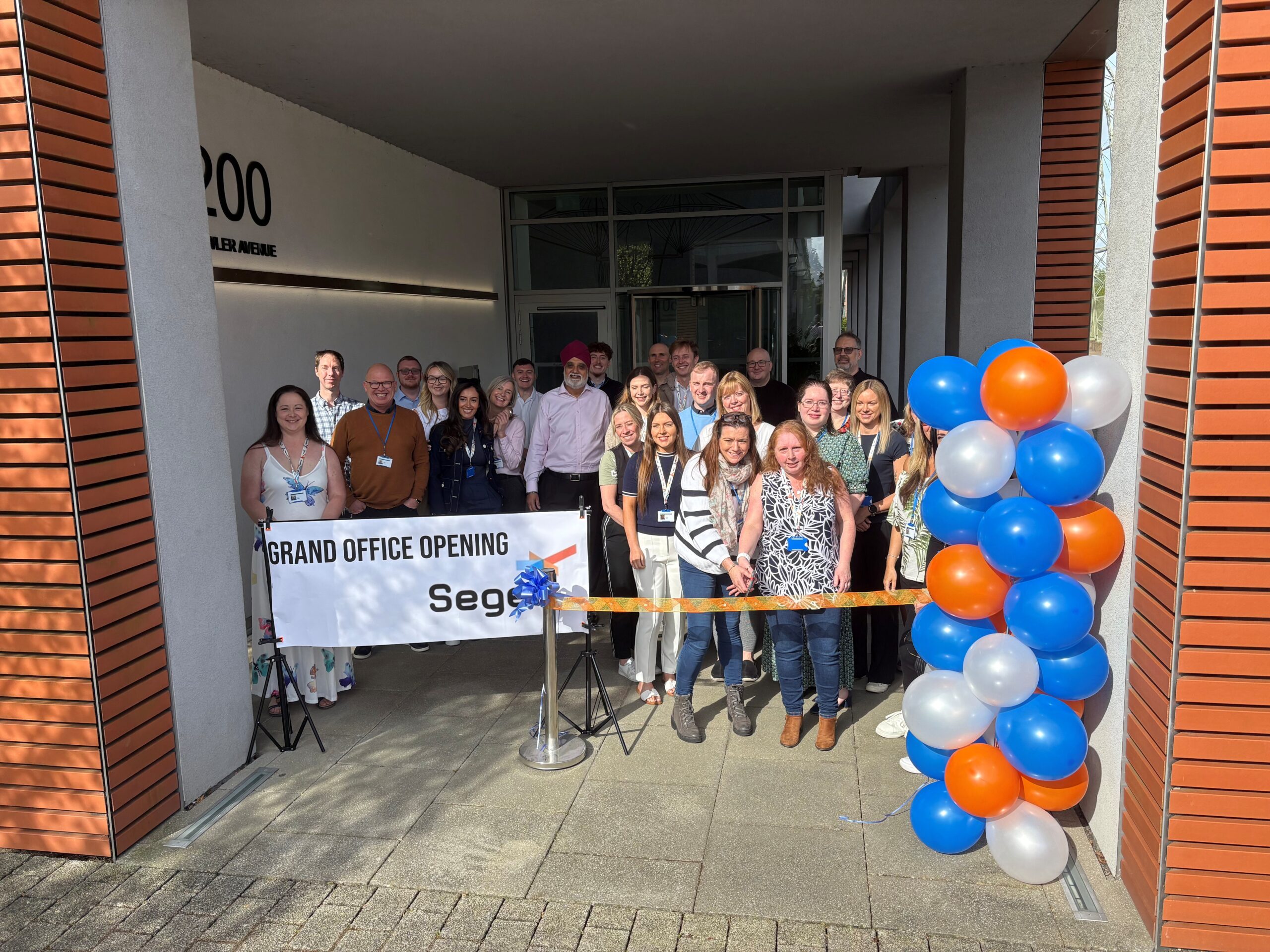 Group of people standing outside a building entrance with a "Grand Office Opening" banner and balloons.